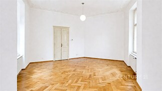 Světlý obývací pokoj s parketovou podlahou a velkými okny, bílé stěny a dekorativní stropní lišty, moderní vzhled a prostorný interiér.