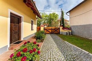 Venkovní příjezdová cesta s dlažbou, vedle udržovaná zahrada s květinami, dřevěná brána a žlutá fasáda staršího rodinného domu.