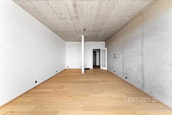 Prázdný obývací pokoj s betonovým stropem, parketovou podlahou a bílými stěnami, nabízí moderní styl a připravenost k osobnímu uzpůsobení.