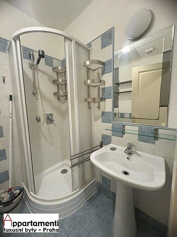 Koupelna s moderním sprchovým koutem, světlé dlaždičky a elegantní umyvadlo, dekorativní dlažba na podlaze a praktické poličky na stěnách.