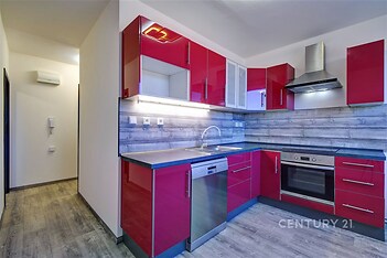 Moderní kuchyň s parketovou podlahou, červenými lesklými skříňkami a vestavěnými spotřebiči, včetně myčky a trouby, nabízí svěží design.