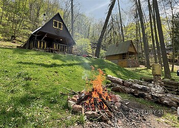 Dřevěná chata s terasou, obklopená lesem, ve svažitém terénu. V popředí je ohniště, ideální pro rekreaci a útěk do přírody.
