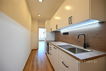 Moderní kuchyň s dřevěnou podlahou, bílými skříňkami a vestavěným osvětlením. Úzký prostor nabízí praktický a stylový design.