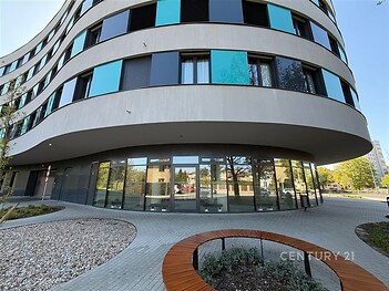 Moderní bytový dům s velkými okny a elegantní fasádou, obklopený dlažbou a upravenými venkovními prostory v klidné části města.