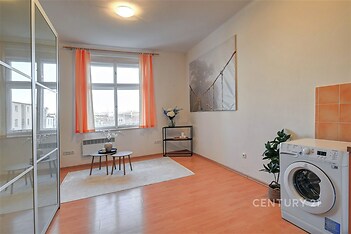 Prostorný obývací pokoj s laminátovou podlahou, velkým oknem a moderním dekorativním prvkem. Světlý interiér s oranžovými závěsy.