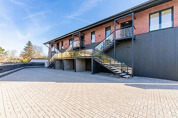 Moderní fasáda bytového domu s cihlovým obkladem, velkými okny a venkovním schodištěm, prostorný dlážděný dvůr s moderním designem.