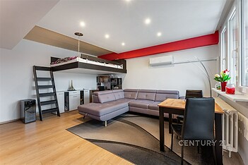 Světlý obývací pokoj s parketovou podlahou, velká okna, moderní klimatizace a podkrovní lůžko vytvářejí útulný a funkční prostor.