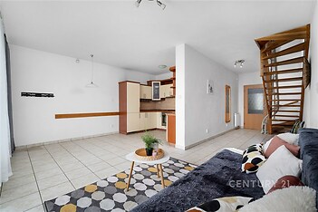 Obývací pokoj s moderní dlažbou, dřevěným schodištěm a světlou kuchyní. Velká okna umožňují přirozené světlo a vytváří prostorný dojem.