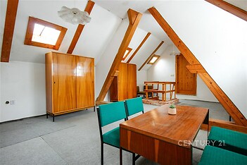 Podkrovní pokoj s dřevěnými trámy, vestavěnou skříní a světlými stěnami, ideální pro kreativní úpravy, nabízí útulný prostor s přirozeným světlem.