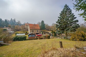Rodinný dům s červenou střechou na rozlehlém pozemku, obklopený zelení a vzrostlými stromy, ideální pro klidné bydlení na okraji města.