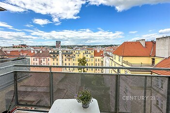 Balkon s kovovým zábradlím a výhledem na město, světlé cihlové budovy s červenými střechami, modrá obloha s mraky
