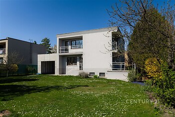 Moderní rodinný dům s bílou fasádou a velkými okny, nacházející se uprostřed prostorné zahrady s udržovaným trávníkem.