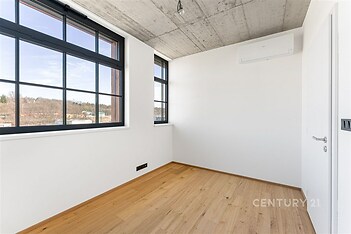 Útulná místnost s parketovou podlahou, velkými okny a moderními prvky, jako je betonový strop a bílé stěny, nabízí světlý prostor.