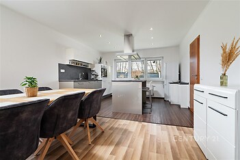 Moderní kuchyň s parketovou podlahou, velkými okny a vestavěnými spotřebiči. Světlý otevřený prostor s dřevěným jídelním stolem a světlým interiérem.