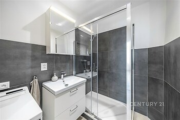 Moderní koupelna s prostorným sprchovým koutem, šedými obklady a elegantním nábytkem. Velké zrcadlo a čistý design vytvářejí světlý prostor.