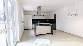 Moderní kuchyň s dlažbou, velká okna zajišťující světlost, elegantní černobílá linka s centrálním ostrůvkem, závěsná svítidla přidávají styl.