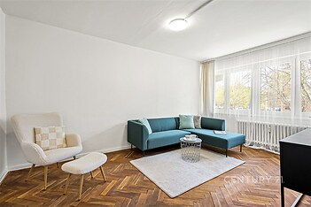 Světlý obývací pokoj s parketovou podlahou, velká okna zabezpečující dostatek světla, moderní nábytek a světlé stěny.