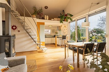 Světlý obývací pokoj s dřevěnou podlahou a velkými okny propojený s moderní kuchyní, krbová kamna vytvářejí útulnou atmosféru.