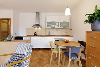 Kuchyň s parketovou podlahou, moderní bílá linka, vestavěné spotřebiče, světlé dřevo nábytku, kytka na komodě.