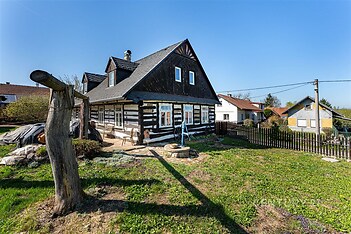 Tradiční roubenka s dřevěnými trámy, oplocený pozemek s udržovanou zahradou, klidné venkovské prostředí.