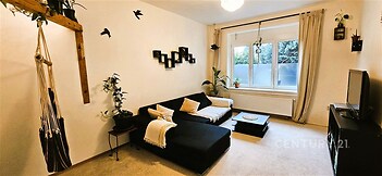 Světlý obývací pokoj s moderní výzdobou, kobercovou podlahou, velkými okny a minimalistickým nábytkem vytváří útulný prostor pro relaxaci.