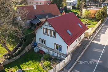 Dvoupodlažní rodinný dům s červenou střechou, zahradou a dřevěným oplocením, nacházející se v klidné rezidenční čtvrti.
