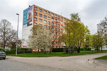 Vícepatrový bytový dům s moderní fasádou, obklopený zelení, stromy a parkovými plochami, vytváří příjemné městské prostředí.