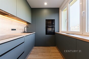 Moderní kuchyň s parketovou podlahou a vestavěnými spotřebiči, velká okna poskytují dostatek světla, elegantní a funkční design.