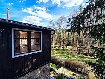 Výhled z dřevěné chaty s kamennou podezdívkou na udržovanou zahradu obklopenou stromy, přírodní materiály a malebná lokalita