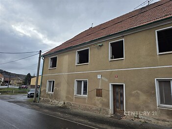 Prázdný dvoupatrový dům s šedou omítkou a červenou střechou, vhodný k rekonstrukci, nacházející se na klidné okrajové ulici.