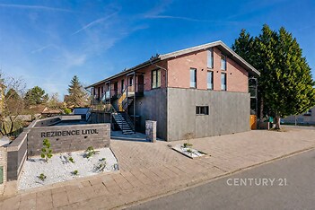 Rezidenční budova s moderní fasádou, velkými okny a cihlovým obkladem. Klidná lokalita s upraveným okolím a přístupovým chodníkem.