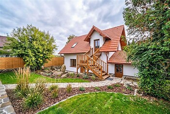 Renovovaný rodinný dům s červenou střechou a dřevěným schodištěm, obklopený upravenou zahradou s kamennou cestičkou, plotem a zelení.