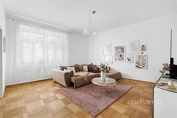 Světlý obývací pokoj s parketovou podlahou, moderní dekorací a velkými okny, poskytující prostorný a útulný interiér.