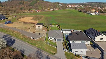 Pohled na moderní rodinný dům s velkými okny a zahradou, nacházející se v klidné oblasti s výhledem na louku a okolní krajinu.