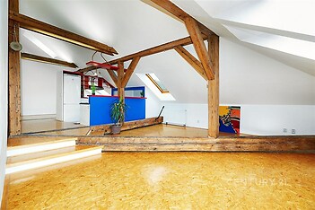 Podkrovní prostor s dřevěnými trámy, parketovou podlahou a moderními prvky, nabízí světlý interiér s vestavěnými doplňky a uměleckým akcentem.
