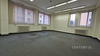 Prostorná kancelář s kobercovou podlahou, velkými okny a vertikálními žaluziemi, vhodná pro moderní pracovní prostředí.