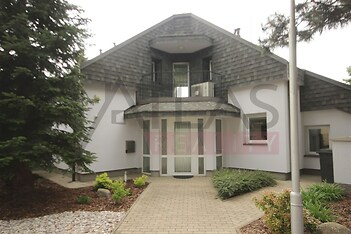 Vchod do rodinného domu s moderní fasádou, střešní tašky a dlážděná cesta, obklopený udržovanou zahradou a jehličnanem.