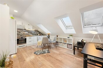 Podkrovní kuchyň a jídelna s moderními spotřebiči, dřevěnou podlahou a velkými střešními okny, vytvářející světlý prostor.