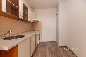 Úzká kuchyň s parketovou podlahou, dřevěnými skříňkami a světlou pracovní deskou, moderní keramická dlažba na zdi kolem sporáku.