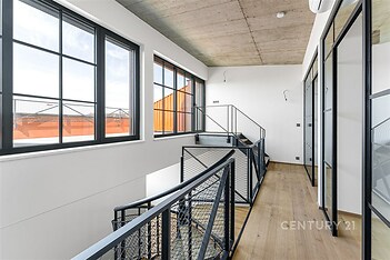 Světlá chodba s velkými okny, moderním průmyslovým stylem a dřevěnou podlahou. Skleněné stěny s kovovými rámy dodávají industriální vzhled.