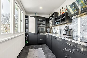 Moderní kuchyň s dlažbou, vestavěnými spotřebiči a velkými okny. Černé skříňky s kovovými úchyty dodávají elegantní vzhled.