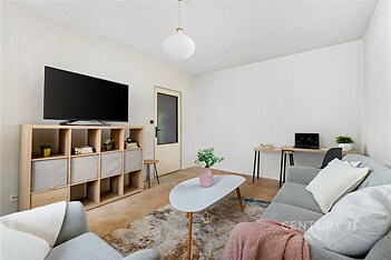 Světlý obývací pokoj s parketovou podlahou, moderním nábytkem a velkým oknem, poskytující útulný prostor pro relaxaci a práci.