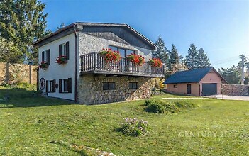 Dvoupodlažní dům na velkém pozemku, obložený kamenem, s květinovými truhlíky na balkoně, zelený a klidný okolní prostor.