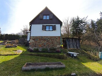 Fasáda venkovského domu s kamennou podezdívkou a dřevěnými obklady, okolní zahrada se vzrostlými stromy a dřevěnou kůlnou.