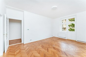 Světlý obývací pokoj s parketovou podlahou, bílými stěnami a velkými okny poskytujícími přirozené světlo, moderní interiér.