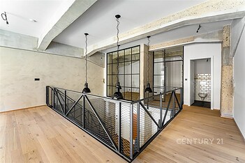 Podkrovní prostor s parketovou podlahou, moderním osvětlením a kovovým zábradlím, kombinace betonu a skla vytváří industriální styl.