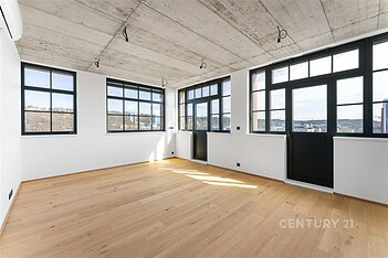 Světlý obývací pokoj s parketovou podlahou a velkými okny, moderní průmyslový styl s betonovým stropem a výhledem na město.