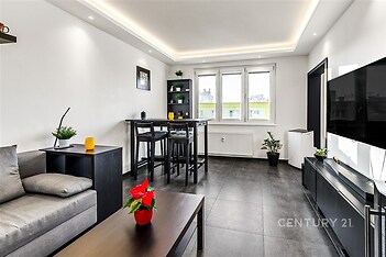 Světlý obývací pokoj s moderní dlažbou, velká okna poskytující dostatek přirozeného světla, elegantní zařízení v rekonstruovaném stylu.