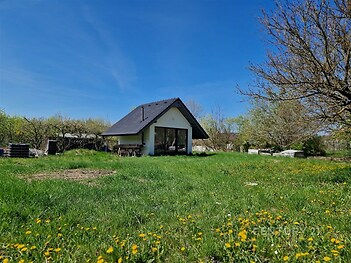 Malý rodinný dům se sedlovou střechou a velkými okny na rozlehlém zeleném pozemku s květinami. Klidné prostředí, ideální pro relaxaci.