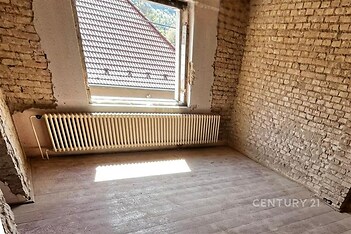 Pokoj ve fázi rekonstrukce s odkrytými cihlovými stěnami, světlým dřevěným podlahovým povrchem a velkým oknem pro přirozené světlo.